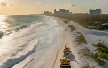 Dos excavadoras amarillas mueven arena en una playa con dunas. El mar con olas está a la izquierda y edificios altos se ven a lo lejos bajo un cielo al atardecer