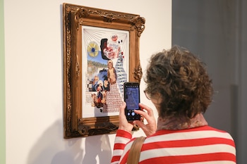 Una mujer sostiene un teléfono móvil frente a un cuadro con marco dorado colgado en una pared blanca