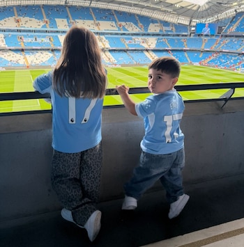 Dos niños con camisetas celestes con el número 17 apoyados en una barandilla mirando una cancha de fútbol