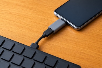 Un teléfono móvil con pantalla apagada conectado a un teclado negro mediante un cable y un adaptador sobre una superficie de madera.