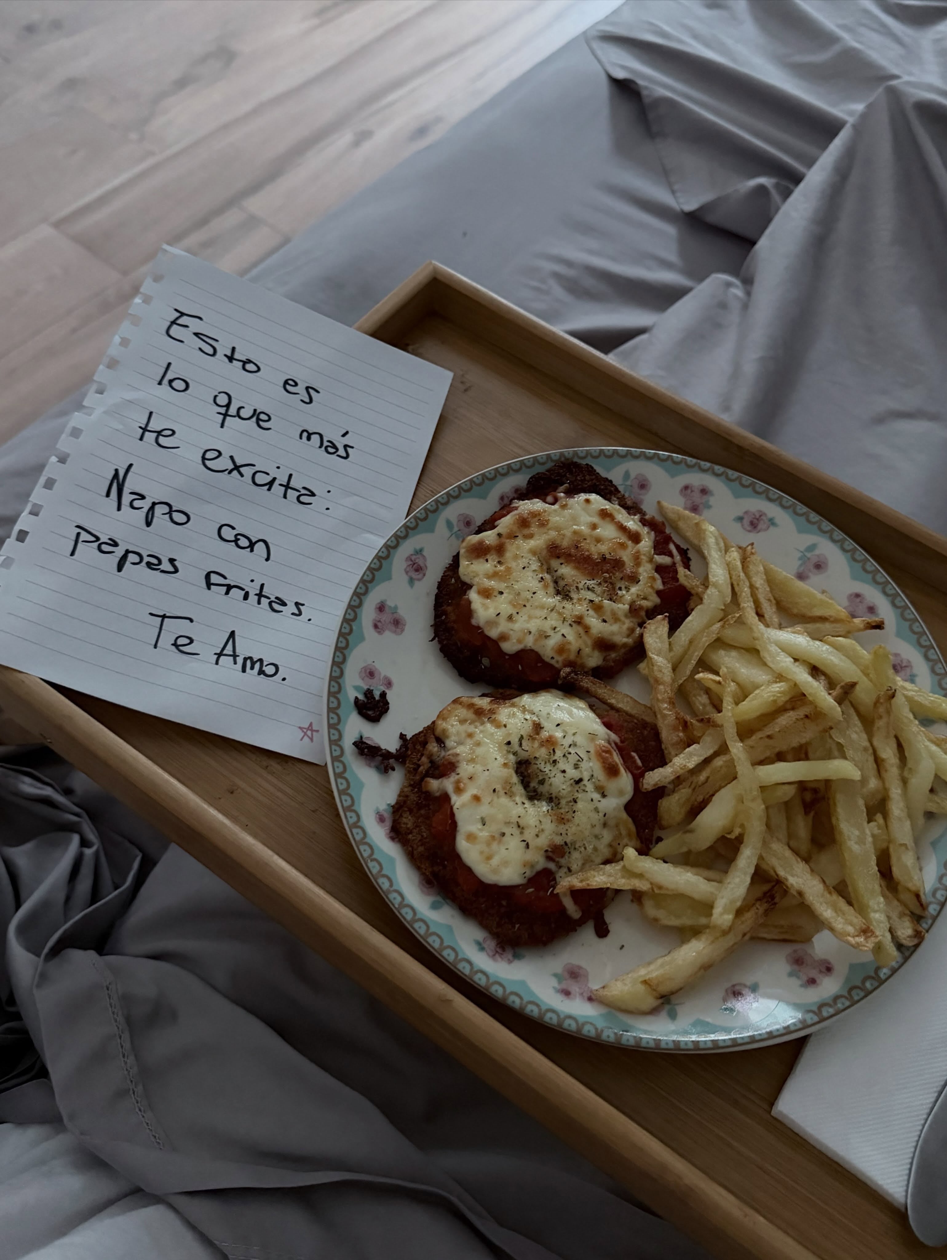 Una bandeja de madera sobre una cama contiene un plato con milanesas a la napolitana, papas fritas y un mensaje manuscrito de afecto.