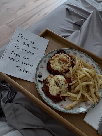 Una bandeja de madera sobre una cama contiene un plato con dos milanesas, papas fritas y una nota manuscrita en un papel de libreta