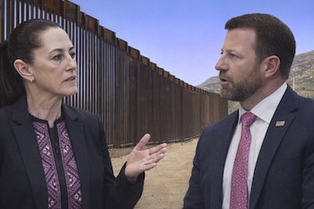 Una mujer y un hombre conversan frente a un muro fronterizo de acero, con el cielo azul y montañas en el desierto como fondo.