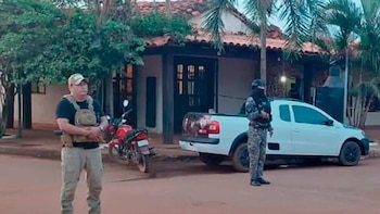 Dos hombres, uno uniformado, se encuentran frente a una casa con techo de tejas. Hay una camioneta blanca, una motocicleta roja y árboles frondosos en la escena