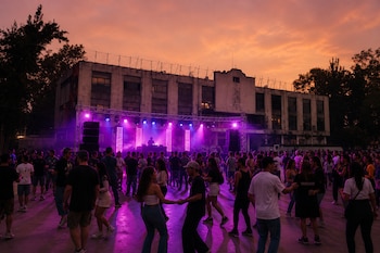 Personas reunidas bailan en una explanada frente a una tarima con luces violetas y un edificio al fondo bajo el cielo del atardecer.