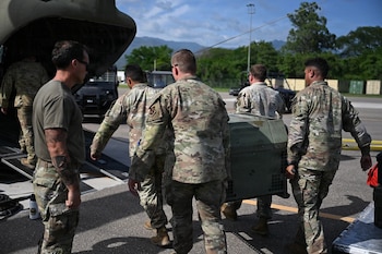 Un grupo de seis militares con uniforme camuflado moviliza una caja metálica verde en una pista de aterrizaje junto a la rampa de una aeronave militar