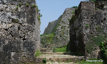 Muros de piedra antiguos cubiertos de vegetación, un sendero de piedra y escalones. Al fondo, más muros de piedra y cielo azul