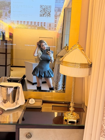 Mujer rubia con vestido gris se fotografía frente a un espejo junto a una lámpara dorada y una hielera metálica sobre una mesa