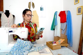 Mujer con gafas y camisa colorida sentada frente a una máquina de coser blanca, sostiene un teléfono celular, con telas y un maniquí
