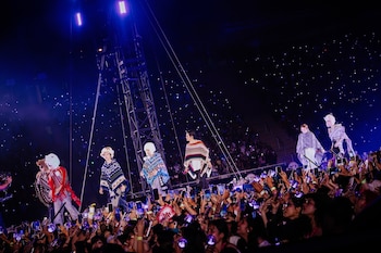 Integrantes del grupo BTS cantan en un escenario iluminado usando ponchos tradicionales frente al público que sostiene teléfonos