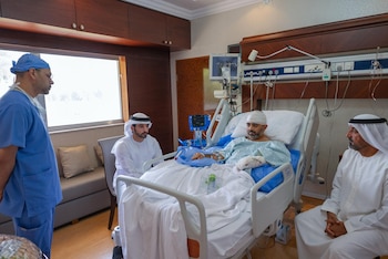 Un paciente vendado en una cama de hospital conversa con tres hombres, uno en ropa médica azul y dos en vestimenta tradicional árabe
