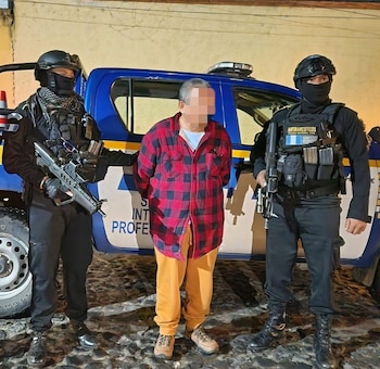Un hombre con camisa a cuadros está escoltado por dos agentes de policía armados y encapuchados junto a un vehículo policial