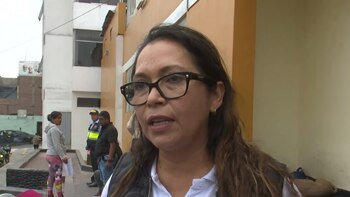 Primer plano de una mujer con gafas oscuras y cabello castaño oscuro, hablando. Detrás, personas, incluyendo un agente de policía, y edificios