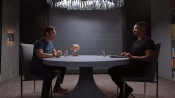 Dos hombres sentados uno frente al otro en una mesa redonda de color claro, con un modelo de cerebro humano y dos tazas metálicas entre ellos