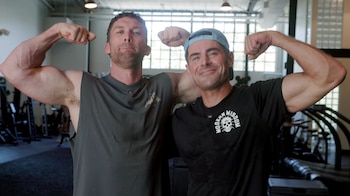 Dos hombres con ropa deportiva flexionan sus bíceps dentro de un gimnasio con máquinas de ejercicio al fondo