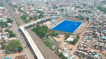 Puente ferroviario de cemento y metal con giro de 90 grados sobre una calle con vehículos, un tren rojo en vías inferiores y personas