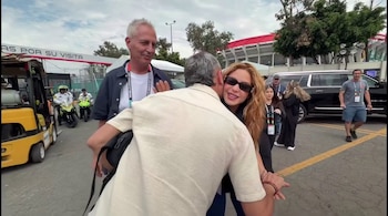 Shakira con gafas de sol abraza a un hombre en primer plano. Marley sonríe detrás. En el fondo, un coche negro, una grúa y dos motocicletas