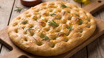 Focaccia redonda con sal gruesa y hojas de romero sobre una tabla de madera en una mesa.
