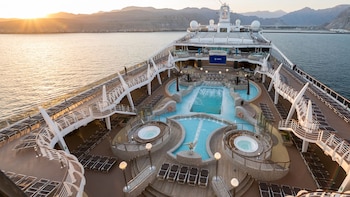 Vista aérea de la cubierta superior del crucero MSC Splendida con piscinas, jacuzzis y tumbonas, bajo un cielo de atardecer y montañas