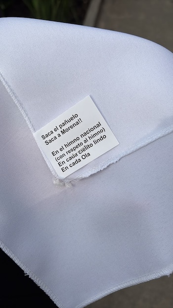 Primer plano de un pañuelo blanco de tela lisa con una etiqueta blanca cosida que lee 'Saca el pañuelo, Saca a Morena!!' en español
