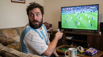 Hombre barbudo con camiseta de Argentina mira a un lado, sorprendido, mientras ve un partido de fútbol en una Smart TV. Sostiene un control remoto y hay snacks en la mesa.