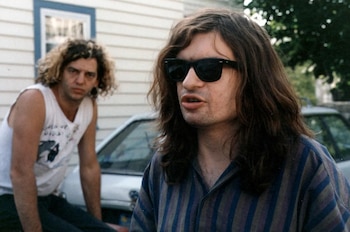 Dos hombres. Uno en primer plano con pelo largo oscuro, gafas de sol y camisa a rayas. El otro, en segundo plano, con pelo rizado claro y camiseta sin mangas. Exterior