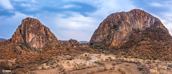 Dos grandes cerros rocosos con un sitio arqueológico y vegetación en el valle central bajo un cielo con nubes
