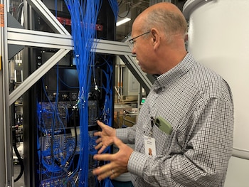 Hombre calvo con gafas y camisa a cuadros gesticula frente a un rack de equipos electrónicos con numerosos cables azules enredados