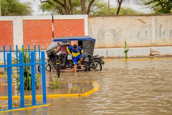 Un escolar descalzo baja de un mototaxi sobre una calle anegada con agua marrón, junto a rejas azules, vegetación y un muro de ladrillos