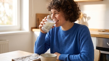 Una mujer de cabello rizado y suéter azul bebe un vaso de leche con semillas de chía sentada junto a una mesa de madera.