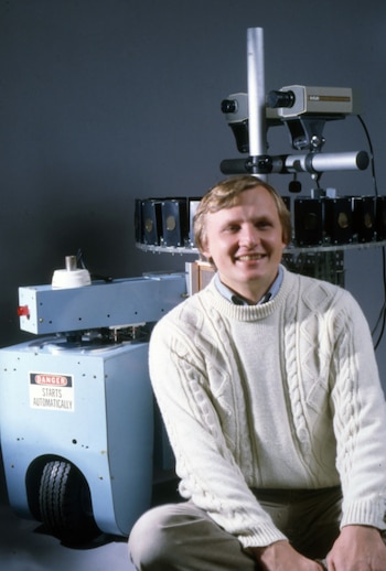 Hombre sonriente sentado sobre un robot con ruedas. Otros dos robots complejos con cámaras y cables en laboratorio. Estanterías, televisores y archivadores en el fondo. Monocromático