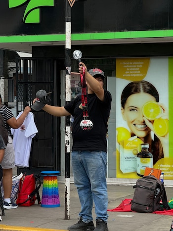 Un hombre sostiene un bastón luminoso y una gorra en la acera, junto a un taburete multicolor y mochilas en la vía pública