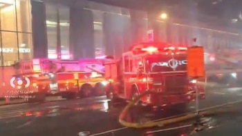 Dos camiones de bomberos rojos con luces de emergencia encendidas y una manguera amarilla en el suelo, con humo o vapor elevándose frente a un edificio
