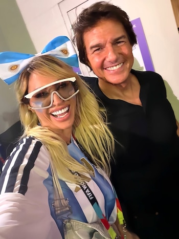 Una mujer rubia con lentes de sol y la camiseta argentina sonríe junto a un hombre con camisa negra, ambos mirando a cámara
