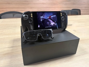 Consola de videojuegos portátil encendida y gafas oscuras sobre una caja negra dispuestas encima de una mesa de madera