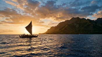 Silueta de una canoa de doble casco con vela triangular navegando en el mar, con montañas tropicales y un cielo al atardecer de fondo.