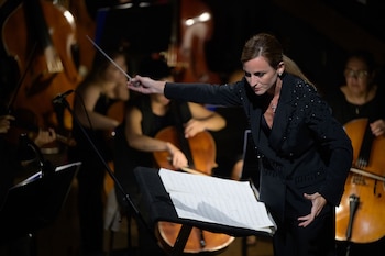 Beatrice Venezi, vestida de negro, dirige una orquesta con batuta y una expresión concentrada, con partituras en un atril y músicos con violonchelos al fondo