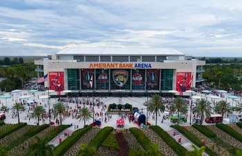 Vista aérea de la Amerant Bank Arena. Multitud de personas en la plaza exterior, con palmeras, zonas verdes, carpas, bandera de Estados Unidos y pancartas deportivas