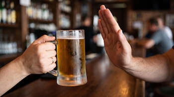 Una mano sostiene una jarra de cerveza helada con condensación, mientras otra mano la rechaza con la palma abierta. Fondo de bar difuminado.