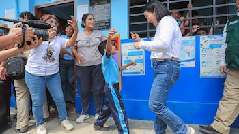 Mujer con camisa blanca y jeans interactúa con niño de uniforme escolar azul. Varias personas adultas y niños detrás de una reja en un edificio azul