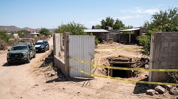 Vehículos de fuerzas de seguridad junto a un portón blanco abierto y una excavación rodeada con cinta de precaución.