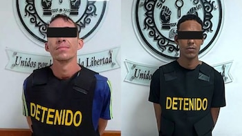 Dos hombres con chalecos negros que llevan la palabra detenido y franjas negras sobre los ojos frente a emblemas policiales en una pared