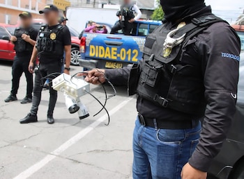 Agente con chaleco táctico sostiene un dispositivo con cámaras y cables, junto a otros efectivos y un vehículo policial