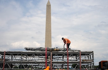 Hombre con camiseta naranja y gorra negra sobre estructura metálica de andamios; detrás, se eleva el Monumento a Washington bajo un cielo nublado