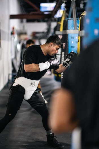 Un hombre con guantes de boxeo negros, protector inguinal blanco y ropa deportiva oscura golpea un saco de boxeo en un gimnasio