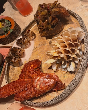 Una bandeja de piedra sostiene cuatro cabezas de piraña, una cola de pescado frito, frutos de achiote y una estructura de conchas