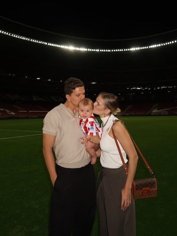 Gianluca Simeone besa la cabeza de su hijo Faustino, mientras Eva Bargiela besa su mejilla, ambos con el bebé en brazos sobre un campo de fútbol iluminado