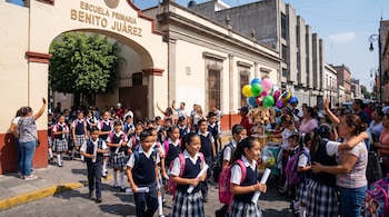 Exterior de escuela primaria Benito Juárez con arco, niños con uniformes a cuadros y mochilas, adultos esperando, globos, vendedores ambulantes en la calle.