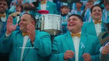 Primer plano de dos hombres sonrientes con trajes turquesa y moños dorados. Uno aplaude, el otro tiene un lazo celeste. Fondo de público y batería