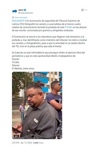 Primer plano de un hombre con barba, gafas y camisa gris sosteniendo un teléfono, con otro hombre joven de camiseta azul detrás y edificios al fondo